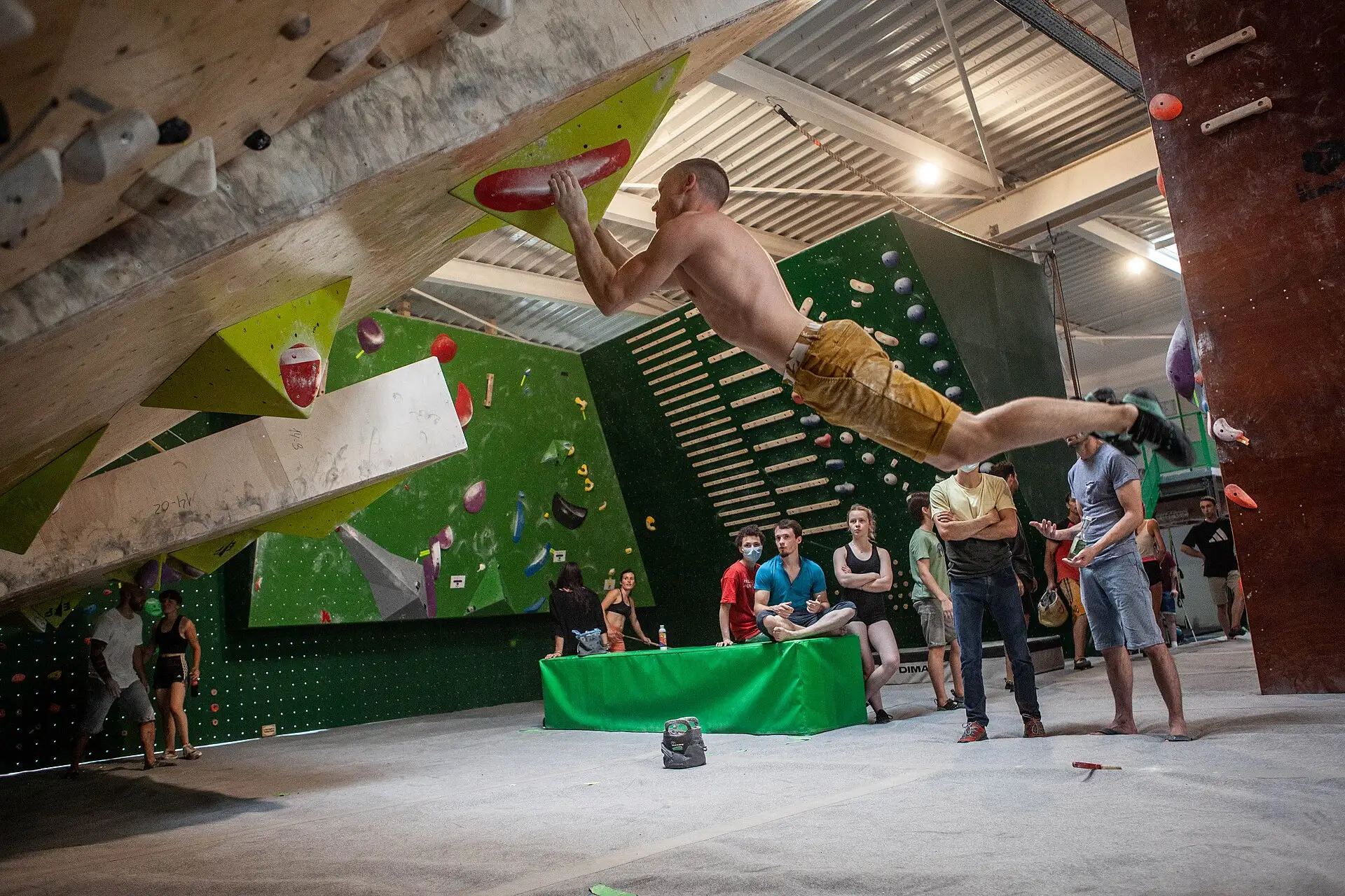 A climber swings feet-off under the steep wooden spray wall at Le Rempart, a bouldering gym, while others watch from the mats.