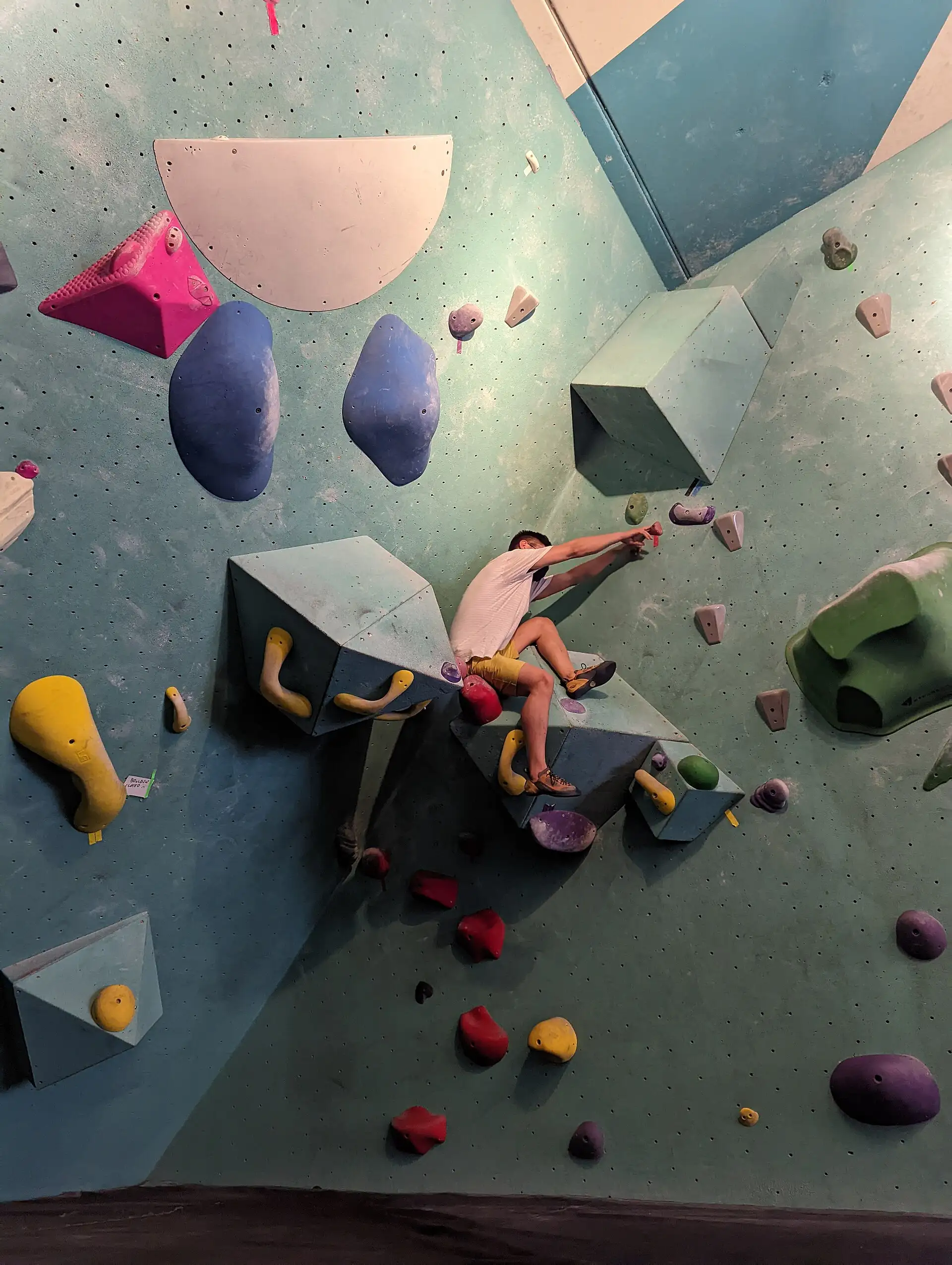 A climber pulls between coloured volumes and holds on an overhanging bouldering wall.