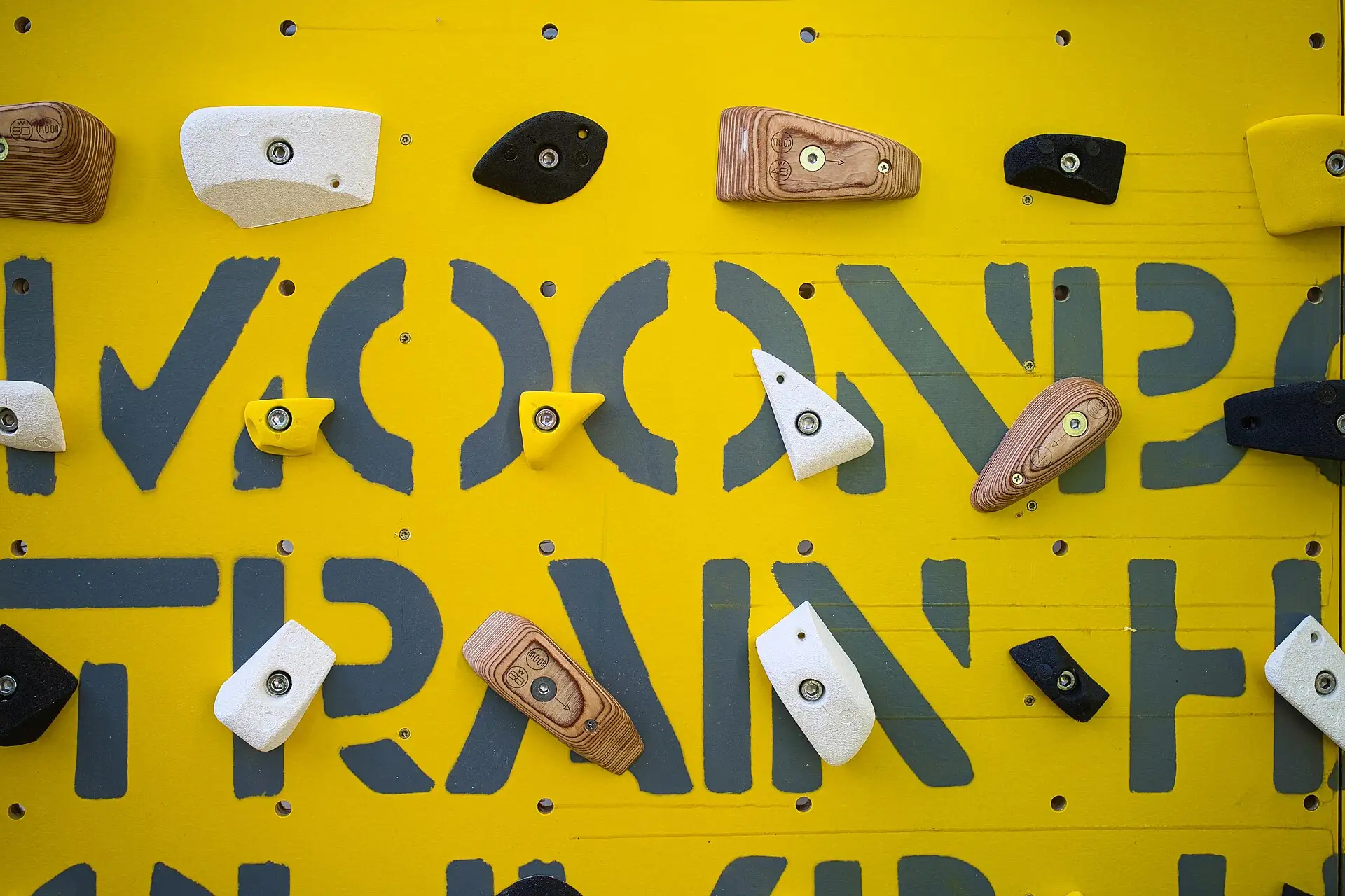 Close up of a yellow MoonBoard panel with wooden, white and black holds bolted over the stenciled MoonBoard lettering.